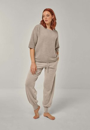 Woman with red hair wearing beige loose knit top and matching drawstring joggers, standing barefoot against plain light background.
