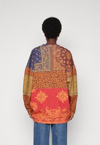 Camisa de manga larga de corte oversized con múltiples patrones: paisley azul, leopardo naranja y floral rojo sobre una base marrón. Textura de tela suave.