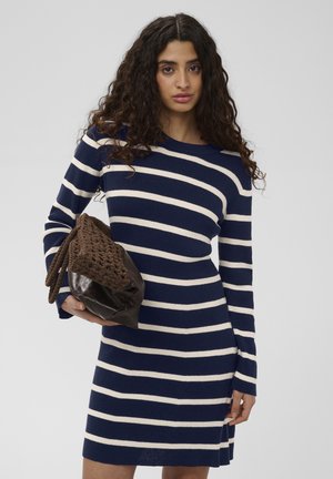 Femme aux longs cheveux bouclés, portant une robe rayée bleu marine et blanche, tenant un grand sac à main marron tressé en cuir.