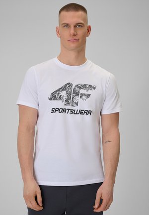 Mężczyzna z krótkimi włosami, ubrany w białą koszulkę 4F Sportswear i ciemne spodnie, stoi na jednolitym szarym tle.