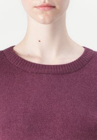 Burgundy stickad tröja med ribbade kanter vid rundhalsen och mjuk textur, med en närbild som framhäver tygets subtila lyster.