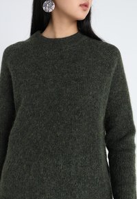 Dunkelgrüner Wollpullover mit Rundhalsausschnitt und langen Ärmeln; weicher, strukturierter Stoff mit etwas lockerer Passform.