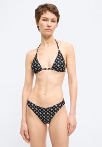 Γυναίκα που φορά μαύρο μπικίνι με μοτίβο και τριγωνικό τοπάκι και σλιπ, στέκεται μπροστά σε απλό φωτεινό φόντο, κοιτάζοντας προς τα εμπρός.