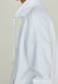 Felpa bianca in pile con colletto alto, chiusura a metà zip e spacco laterale. Presenta una vestibilità comoda e una texture morbida.