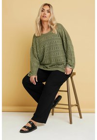 Löst stickad, olivgrön tröja med ögla-mönster, matchad med svarta byxor med vida ben och svarta slide-sandaler med nitar.