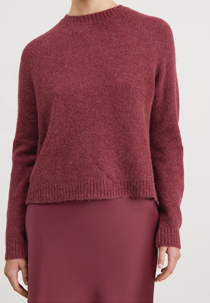 Femme portant un pull bordeaux texturé à manches longues et col rond, associé à une jupe bordeaux lisse, visible des épaules jusqu'aux hanches.