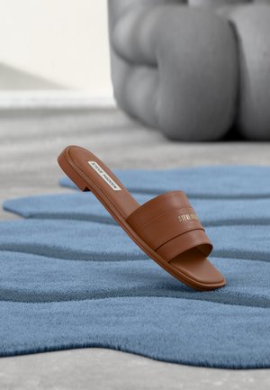 Brązowy skórzany klapek Steve Madden leżący na fakturowanym niebieskim dywanie z rozmytą szarą poduszką w tle.
