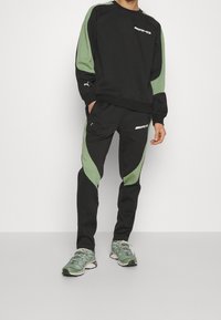 Mężczyzna ubrany w czarny i zielony dres AMG Puma z detalami logo, w połączeniu z zielono-białymi butami sportowymi, stojący na białym tle.