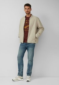 Mężczyzna w beżowej pikowanej kurtce, bordowej bluzie z nadrukiem, niebieskich dżinsach i białych trampkach stoi oparty o białą ścianę.