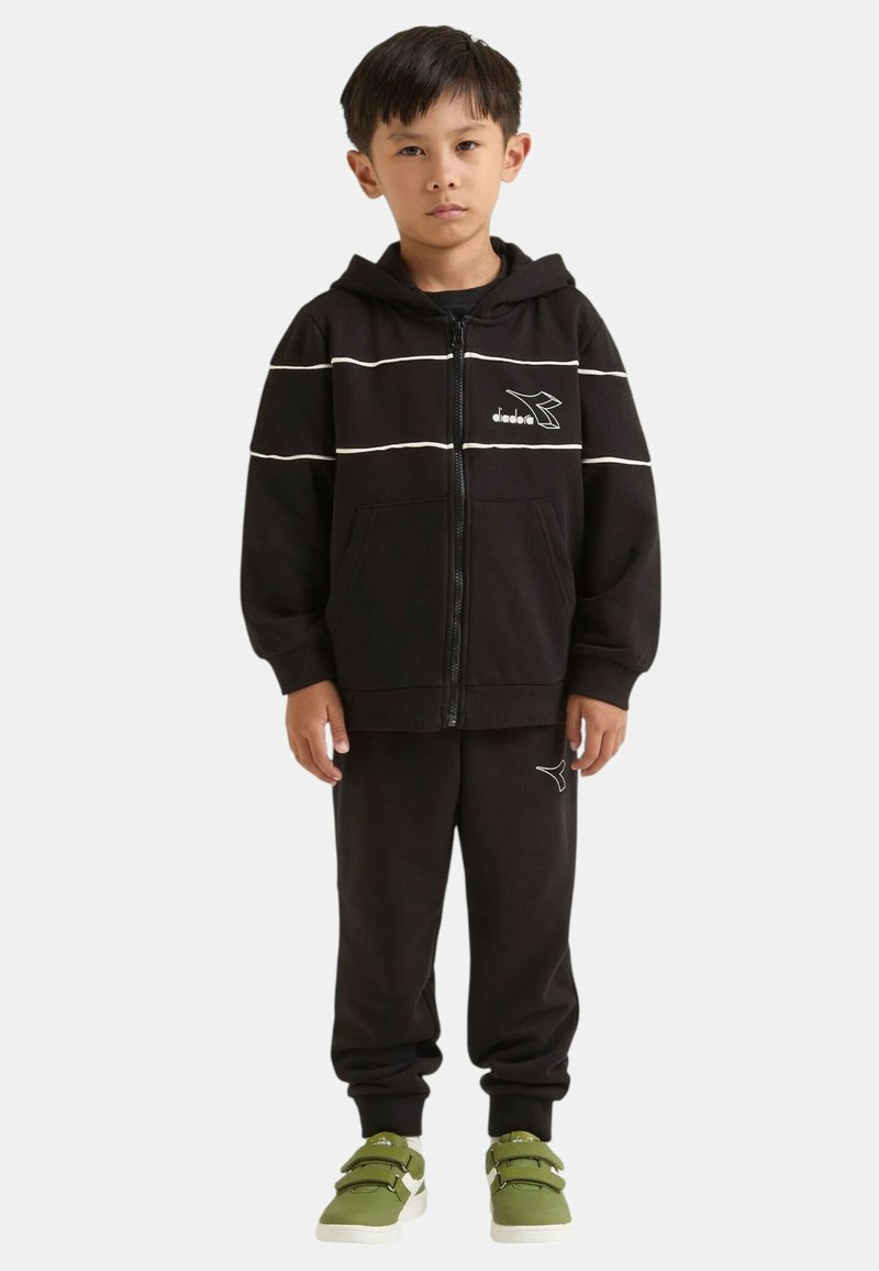 Felpa nera con zip e strisce bianche, con logo, abbinata a pantaloni jogger neri coordinati. Il modello indossa sneakers verdi con doppia fascia.