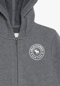 Felpa con zip grigia realizzata in tessuto morbido, caratterizzata da un logo bianco rotondo con un alce e il testo "ABERCROMBIE NEW YORK 1892" sul petto.