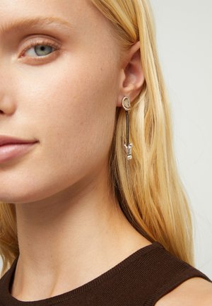 Mujer rubia que lleva una camiseta marrón sin mangas y un pendiente largo de plata y negro con forma de letra Y en la oreja izquierda.