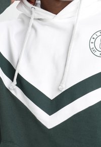 Sudadera con capucha en diseño de bloques de color blanco y verde oscuro, que presenta un patrón de chevron, cordones y un logotipo circular en el pecho.