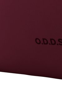 Sac en tissu bordeaux avec une surface texturée, présentant une légère empreinte des lettres "O.D.D.S." dans une teinte plus foncée sur un côté.