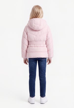 Kind trägt eine hellrosa wattierte Kapuzenjacke, blaue Jeans und weiße Turnschuhe, steht mit dem Rücken zur Kamera vor weißem Hintergrund.
