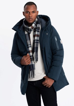 Marineblauer Daunenjacke mit Kapuze und Reißverschluss vorne, kombiniert mit einem karierten Schal in dunklem Blau, Weiß und roten Akzenten. Darunter ein strukturiert gestrickter Pullover.