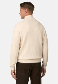 Pull en maille côtelée crème avec un col haut, tissu texturé et ourlet près du corps, associé à un pantalon en velours côtelé marron foncé.