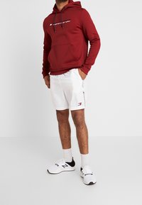 Rode hooded sweatshirt met voorzak en wit logo, gecombineerd met witte shorts met zijbranding. Draagt witte en zwarte sport sneakers.