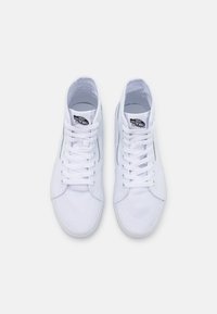 Hvide høj-top sneakers i canvas, med flade snørebånd, forstærket tå og klassisk Vans-logo på tungen, med et slankt, enkelt design.