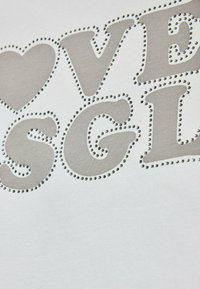 Fehér póló, amelyen egy szürke betűkből készült dizájn látható, szívvel kiegészítve, a betűk körvonalát kis strasszkövek díszítik.