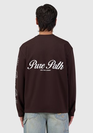 Man facing away wearing a dark long-sleeve shirt with "Pure Path Own Your Journey" text on the back and faded blue jeans.