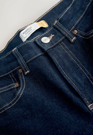 Tumši zili bootcut džinsi ar vidēju jostas augstumu. Iezīmējas ar zelta šuvēm un sudraba pogu aizdari. Etiķete redzama augšpusē.