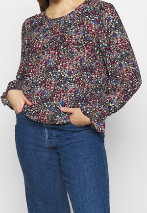 Femme portant un chemisier à manches longues avec un imprimé floral rouge, bleu et blanc, rentrée dans un jean bleu taille haute, les mains dans les poches.