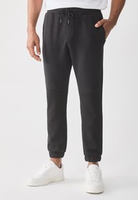 Svarte joggebukser med snøring i livet, sidelommer og elastiske mansjetter, kombinert med hvite joggesko. Slett stoffkvalitet.
