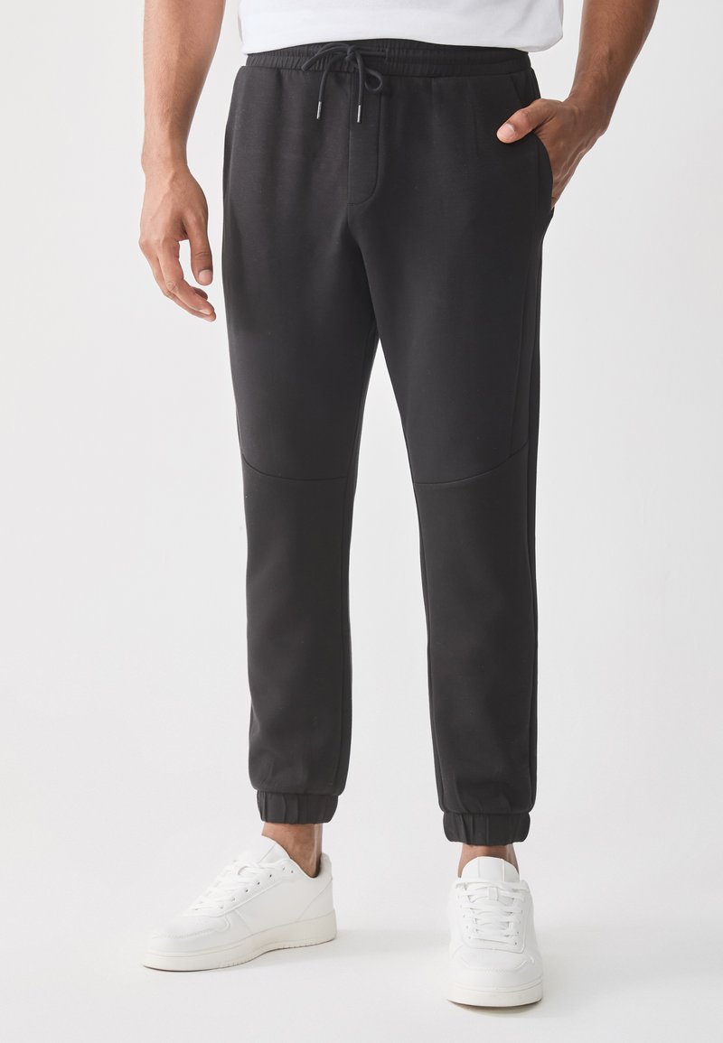 Svarte joggebukser med snøring i livet, sidelommer og elastiske mansjetter, kombinert med hvite joggesko. Slett stoffkvalitet.