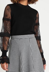 Czarny sweter z dzianiny z przezroczystymi, koronkowymi długimi rękawami oraz koronkowymi detalami przy mankietach, zestawiony z czarno-białą spódnicą w kratkę o wysokim stanie.