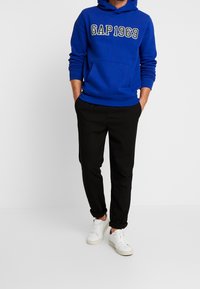 Sudadera azul con "GAP1969" impreso en blanco y negro, combinada con pantalones negros y zapatillas blancas. Presenta un bolsillo canguro en la parte delantera.