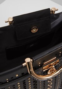 Sac à main noir avec clous dorés, poignée dorée et fermoir, montrant l'intérieur avec une doublure noire et une petite poche à rabat avec un bouton magnétique.