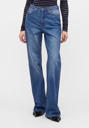 Person iført blå straight-leg jeans, sorte laksko med spids tå og en sort og gråstribet bluse med lange ærmer, stående mod en hvid baggrund.