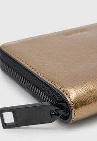 Metalická zlatá peňaženka s čiernym zipsom, ktorá má hladký povrch a značkový ťahák zipsu. Dizajn kombinuje textilné a kožené materiály.