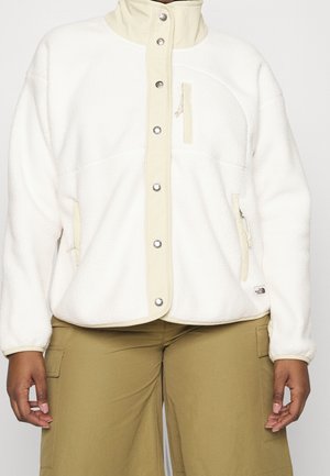 Persona vistiendo una chaqueta de forro polar color crema con botones a presión y bolsillos con cremallera, combinada con pantalones cargo marrones claros.