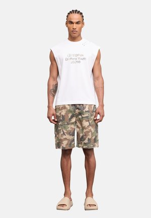Hombre con cabello trenzado, vistiendo una camiseta blanca sin mangas con texto, pantalones cortos de carga de camuflaje y sandalias beige, de pie contra un fondo blanco.