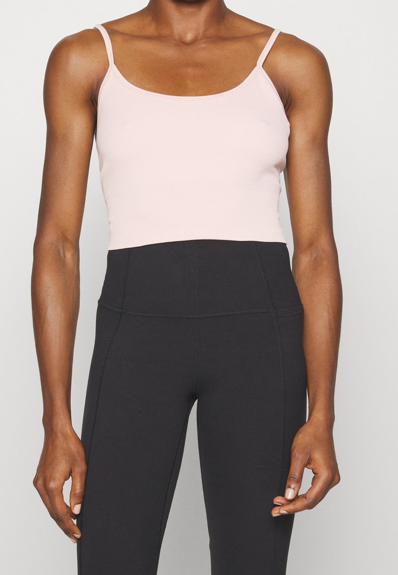 Femme portant un haut court rose clair et un legging noir taille haute, se tenant devant un fond uni.