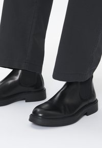 Botas Chelsea de cuero negro con paneles laterales elásticos, suelas planas y puntas redondeadas, combinadas con pantalones negros. Textura suave y diseño minimalista.