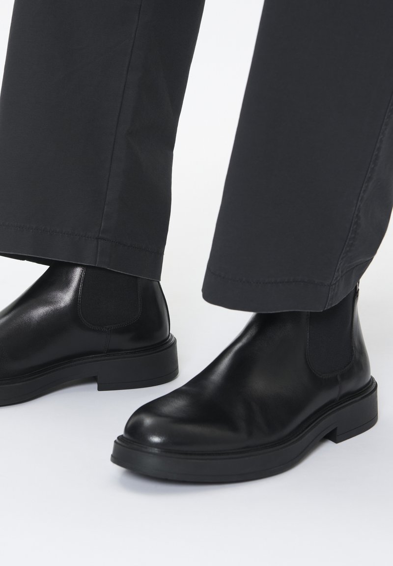 Botas Chelsea de cuero negro con paneles laterales elásticos, suelas planas y puntas redondeadas, combinadas con pantalones negros. Textura suave y diseño minimalista.