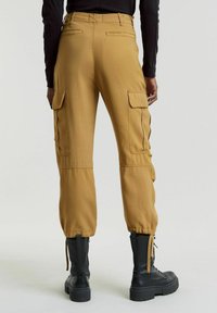 Pantaloni cargo casual in tessuto marrone chiaro con orli regolabili, tasche laterali e un design a gamba dritta, abbinati a stivali da combattimento neri.