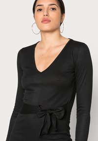 Top preto de manga longa com nervuras e decote em V profundo, apresentando um cinto de tecido atado na cintura. Textura suave e design ajustado.