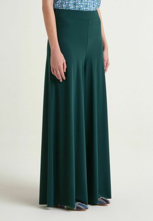 Donna che indossa una gonna a gamba larga verde scuro a vita alta abbinata a scarpe blu con fantasia e una maglietta blu con fantasia infilata dentro.