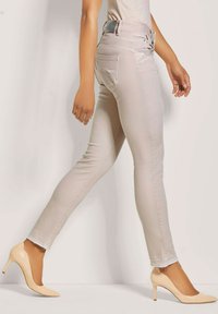 Lichtbeige skinny jeans met een hoge tailleband, subtiele slijtage aan de zoom, gecombineerd met naakthoge hakken, in een wandelhouding geplaatst.