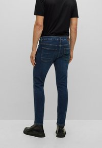 Hombre con jeans oscuros ajustados, camiseta negra y botas de cuero negras, de pie contra un fondo gris claro liso.