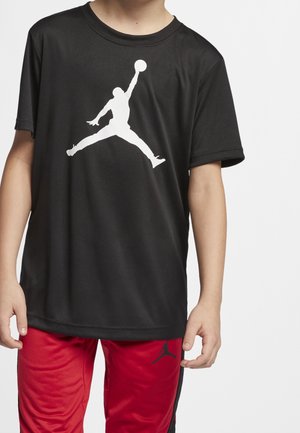 Kind draagt zwart T-shirt met witte silhouet van een basketballer en rode broek met klein zwart logo op de linker dij.