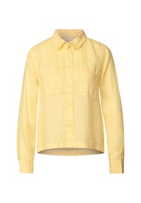 Camicia di lino gialla con colletto a bottoni, maniche lunghe e due tasche sul petto. Orlo leggermente arrotondato. Tessuto textured.