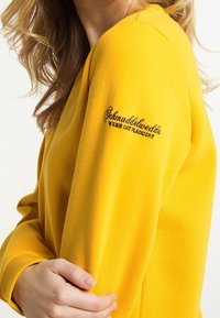Žlutá mikina s volným střihem. Obsahuje žebrované manžety a vyšívaný text v černé barvě na levém rukávu. Měkká textura.