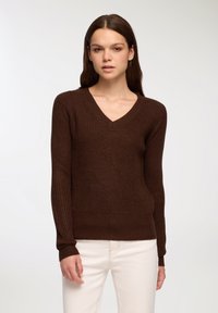 Maglione di lana marrone con scollo a V, polsini e orlo a coste. Tessuto testurizzato, design aderente, indossato con pantaloni chiari.