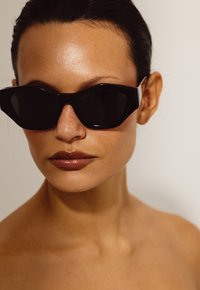 Lunettes de soleil angulaires noires avec une finition brillante, présentant des montures épaisses et des verres foncés. La peau est lisse avec un léger éclat, mettant en valeur des taches de rousseur.