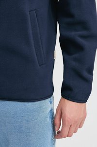 Mornarsko flis jakna z bočno žepe, gladko teksturo, zaobljenim robom in etiketo na spodnjem levem delu. Usklajena s svetlo modrimi jeans hlačami.